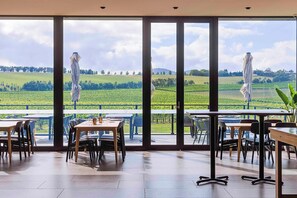 Un restaurant doté de grandes fenêtres donnant sur un paysage verdoyant. Des tables et des chaises sont disposées à l’intérieur.