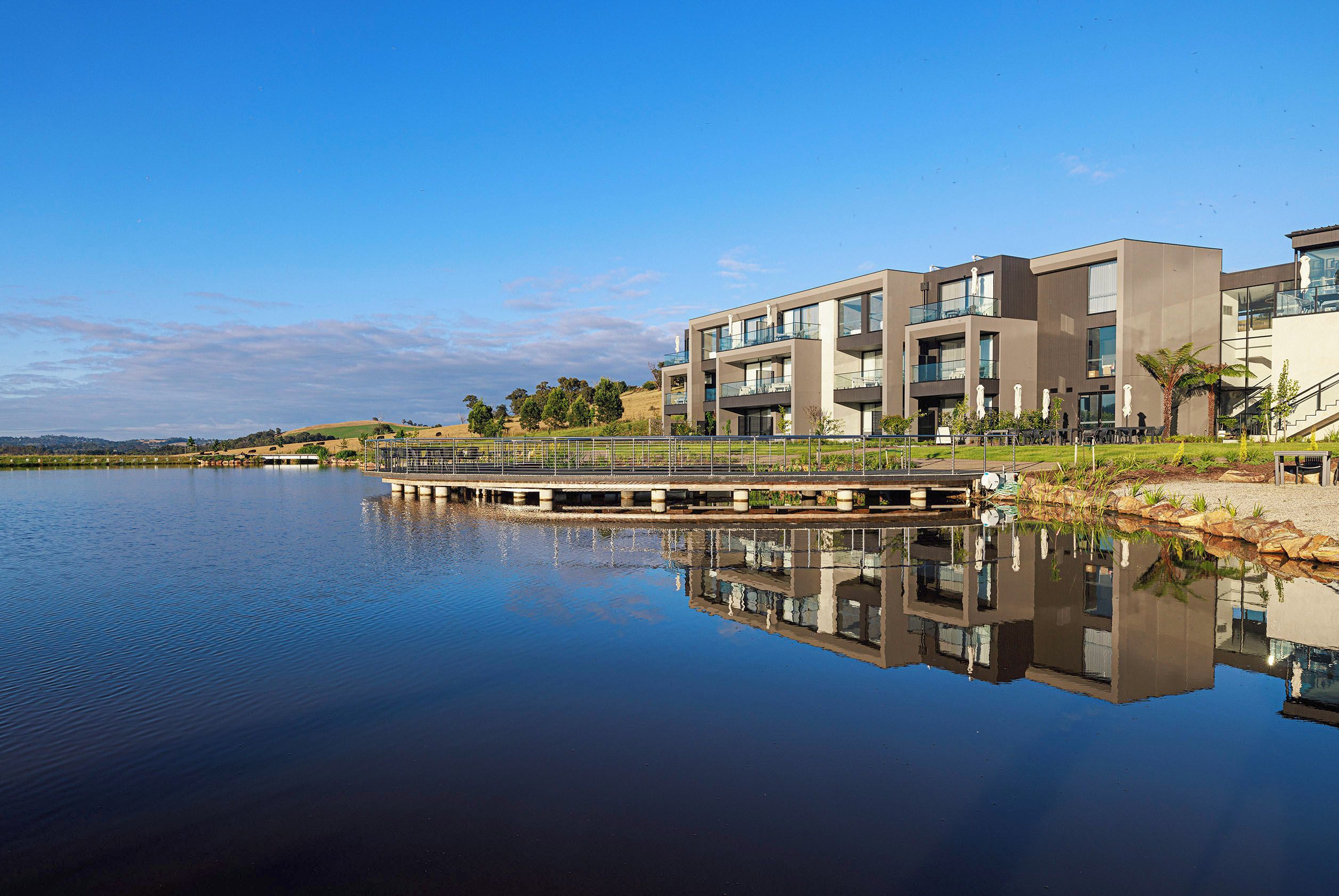 A modern building with a reflection in calm water, a bridge, and a clear sky.