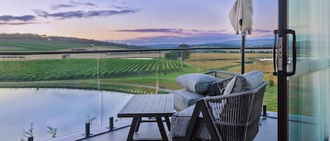 A balcony with a view of a vineyard and a body of water, a wicker chair, and a table.