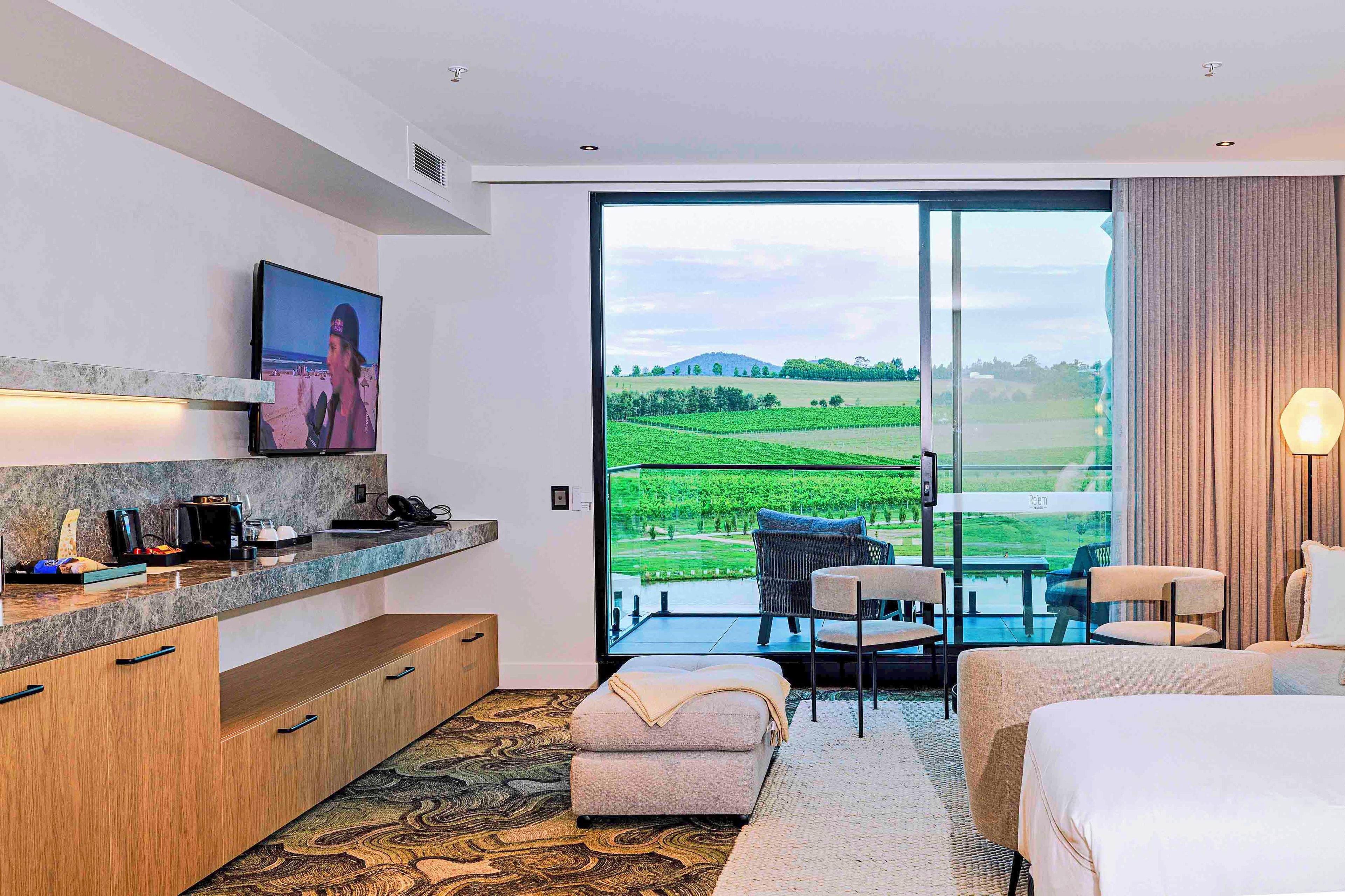 A modern hotel room with a large sliding glass door leading to a balcony, a flat-screen TV mounted on the wall, a small kitchenette, and a cozy seating area with a sofa and chairs.