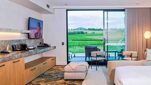 Une chambre d’hôtel moderne dotée d’une grande porte-fenêtre coulissante donnant sur un balcon, d’un téléviseur à écran plat fixé au mur, d’une petite kitchenette et d’un coin salon confortable avec un canapé et des fauteuils.