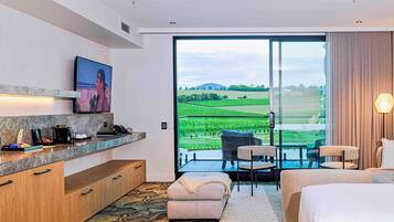 A modern hotel room with a large sliding glass door leading to a balcony, a flat-screen TV mounted on the wall, a small kitchenette, and a cozy seating area with a sofa and chairs.