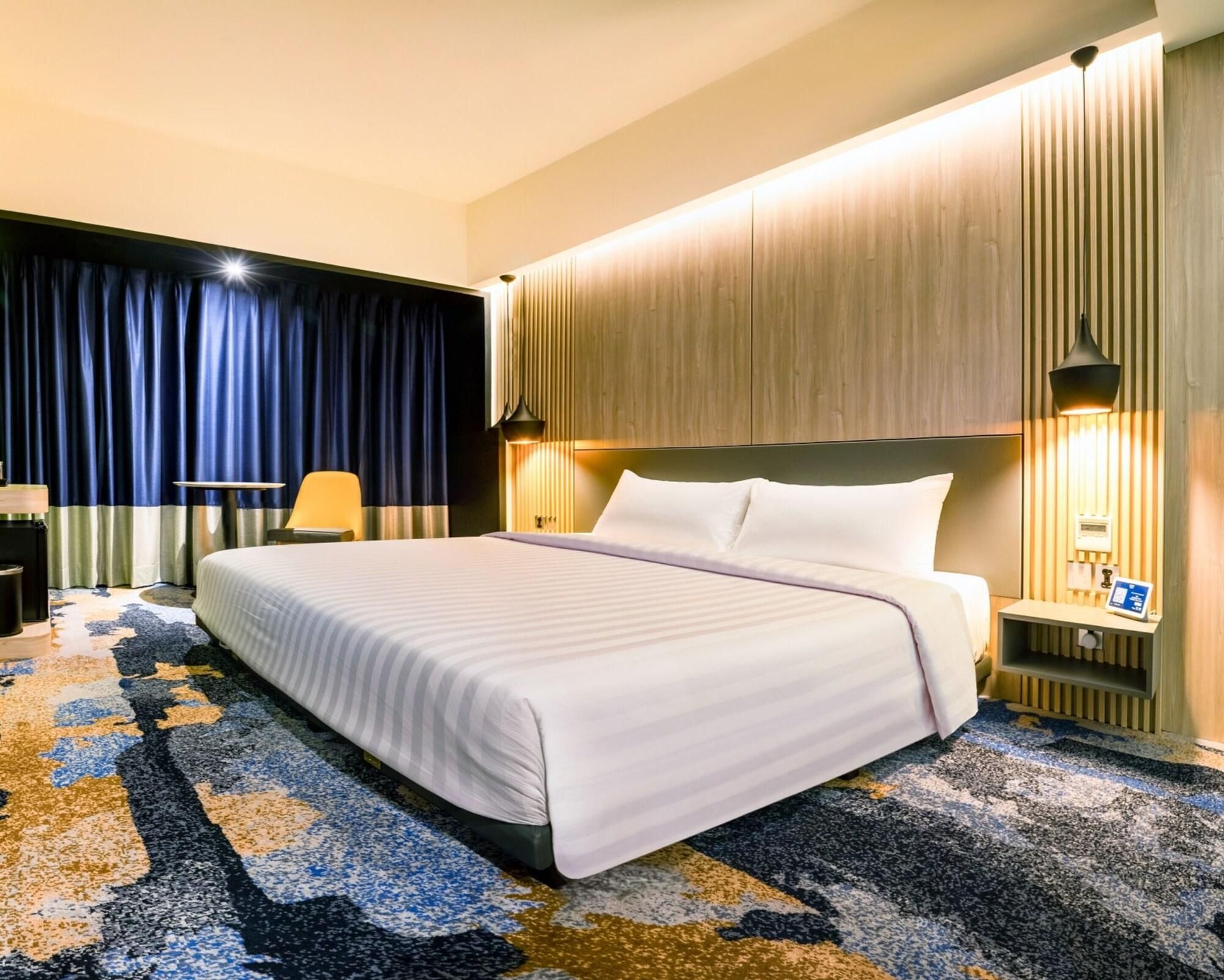A hotel room with a large bed, a desk, a chair, and a nightstand with a lamp.