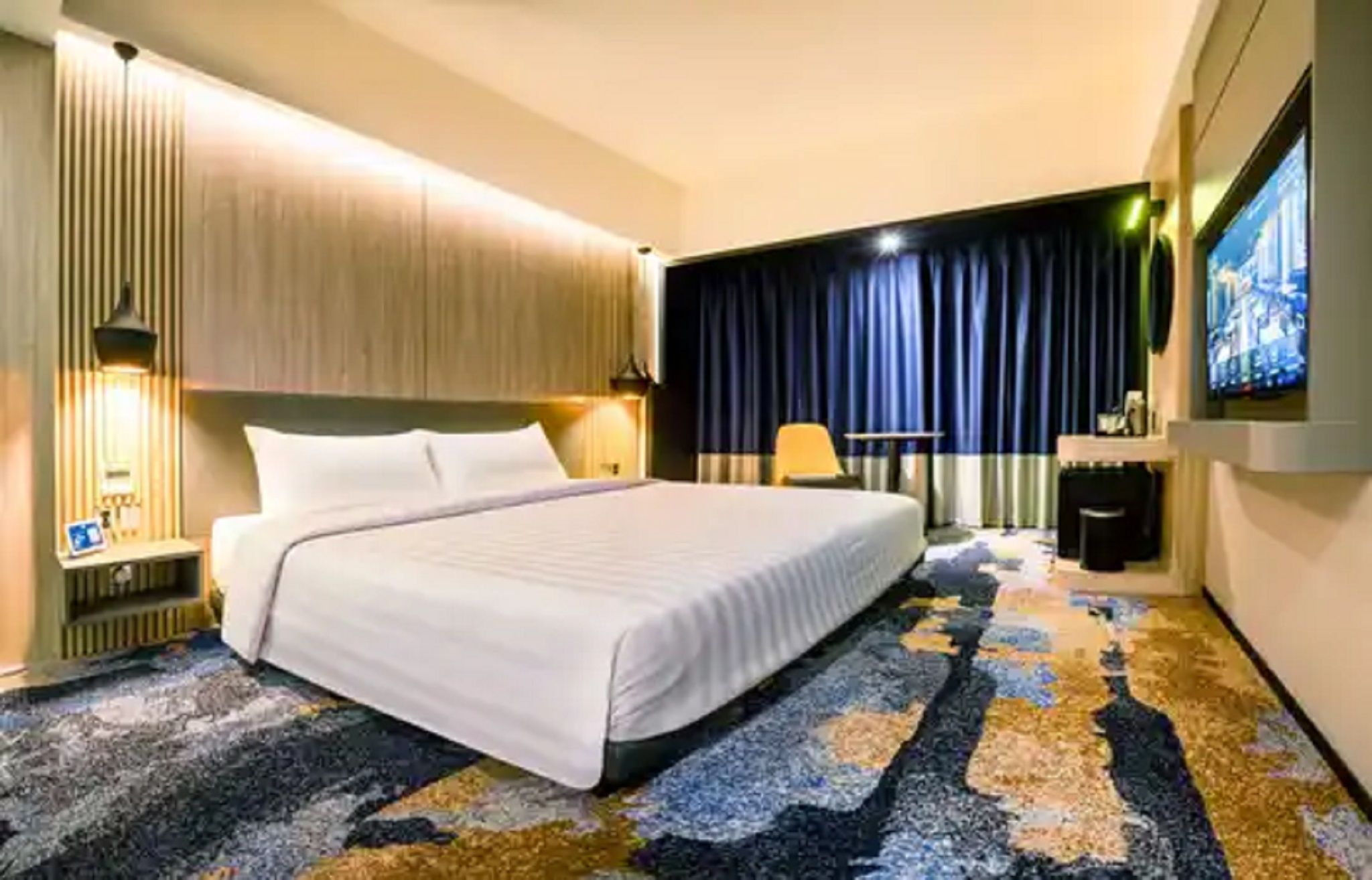 A hotel room with a bed, a desk, a chair, a television, and a patterned carpet.