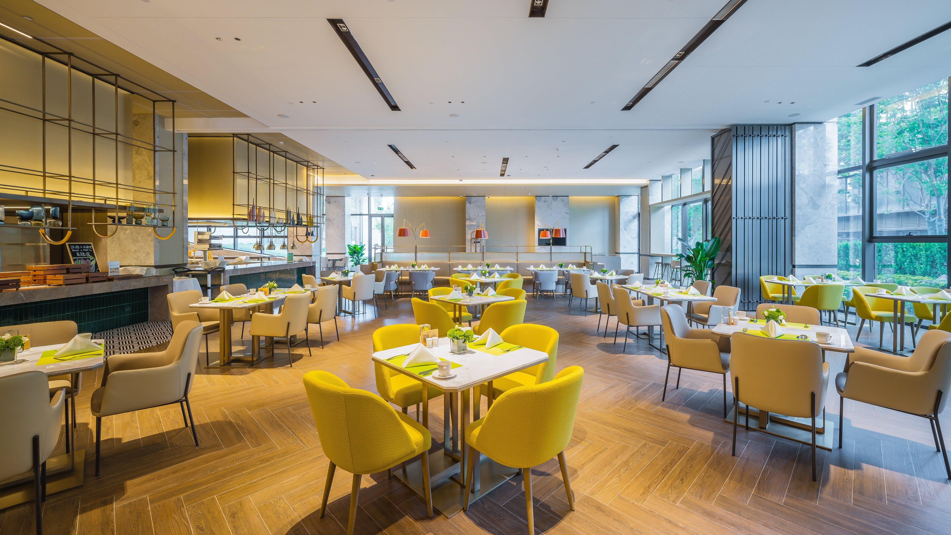 A spacious dining area with multiple tables and chairs, large windows, and a modern kitchen visible in the background.