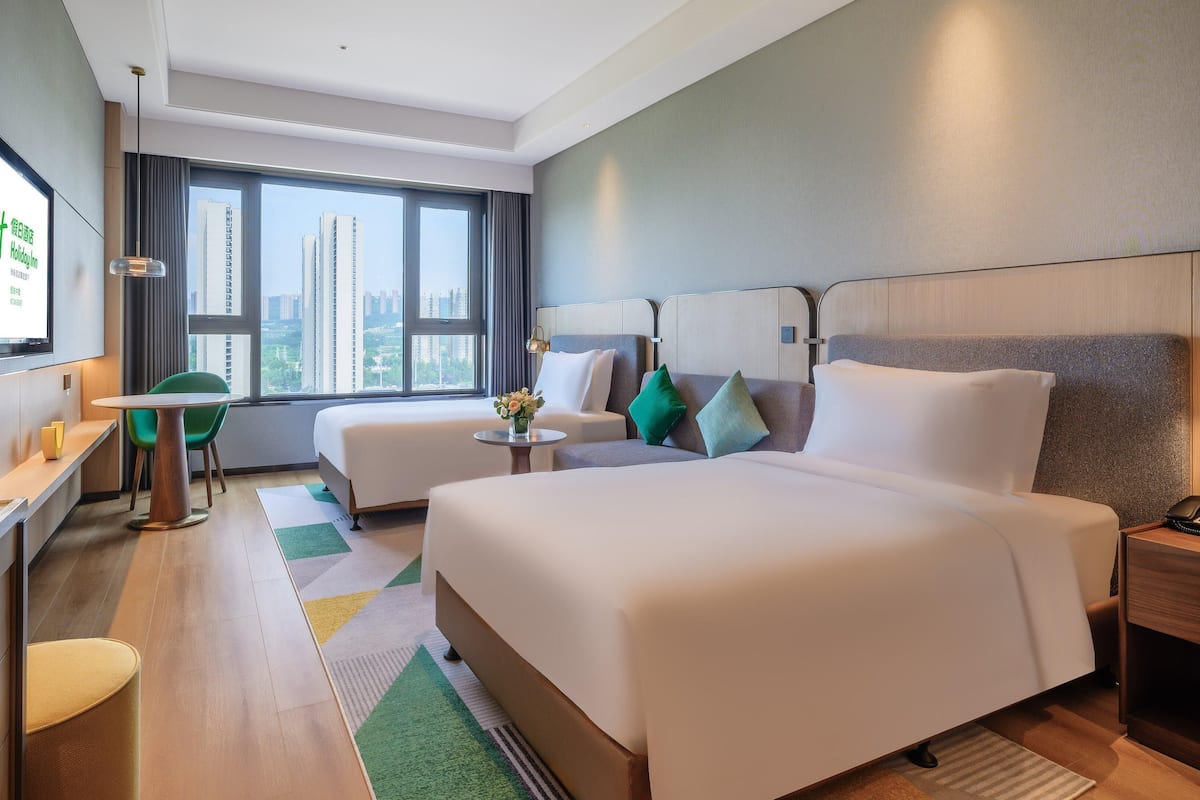 Une chambre d’hôtel équipée d’un lit, d’un bureau, d’une chaise, d’une petite table et offrant une vue sur la ville grâce à de grandes fenêtres.