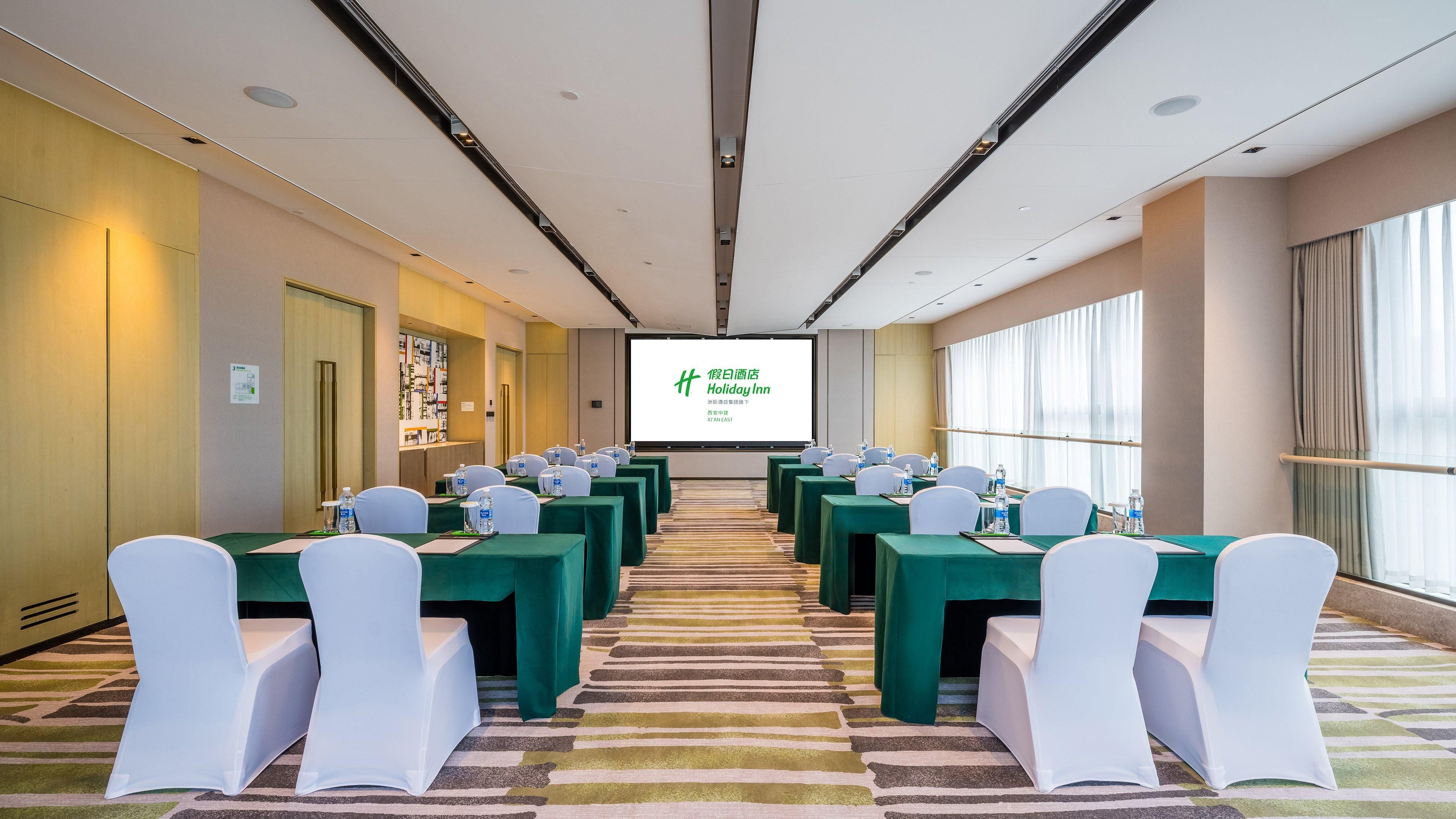 Una sala conferenze con file di tavoli e sedie, uno schermo per proiettore e un'ampia finestra.