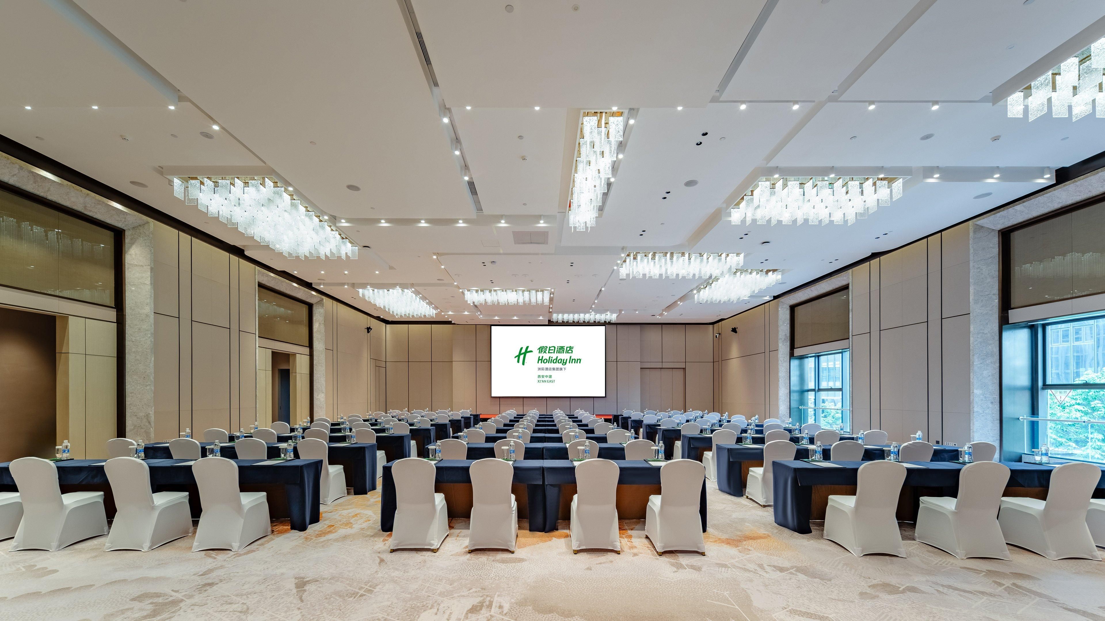 A conference room with rows of tables and chairs, a large screen at the front, and a modern chandelier.