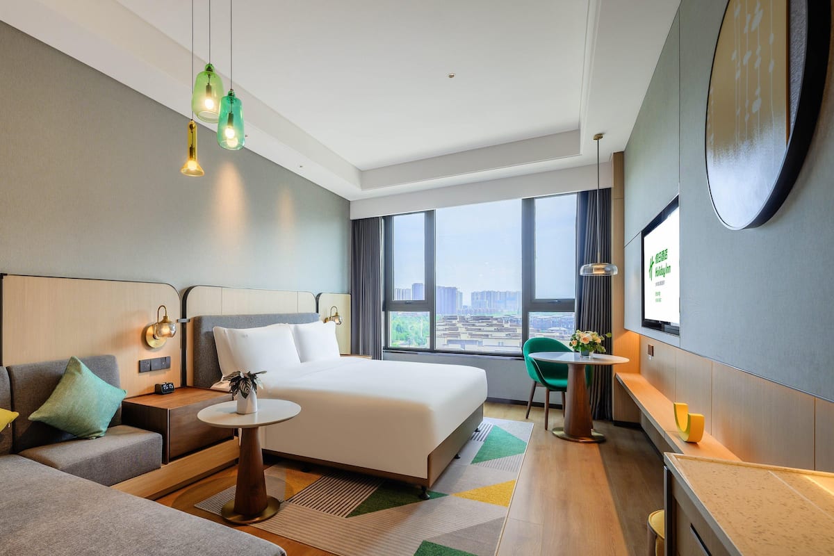 Une chambre d’hôtel moderne, dotée d’un grand lit, d’un canapé, d’une petite table et offrant une vue sur la ville grâce à de grandes fenêtres.
