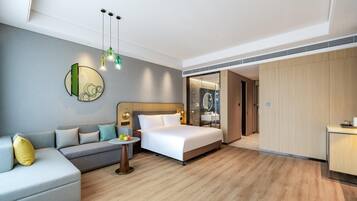 Una habitación de hotel moderna con cama, sofá, una mesita con fruta y un baño visible a través de una puerta abierta.