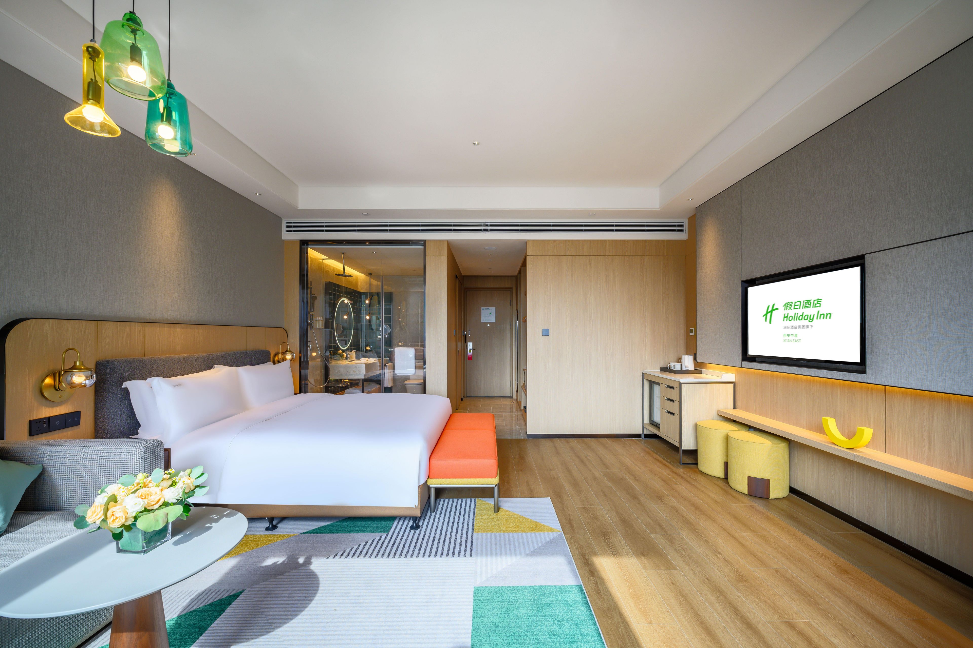 Una habitación de hotel moderna con una cama grande, un escritorio y una pantalla de televisión mostrando información del hotel.