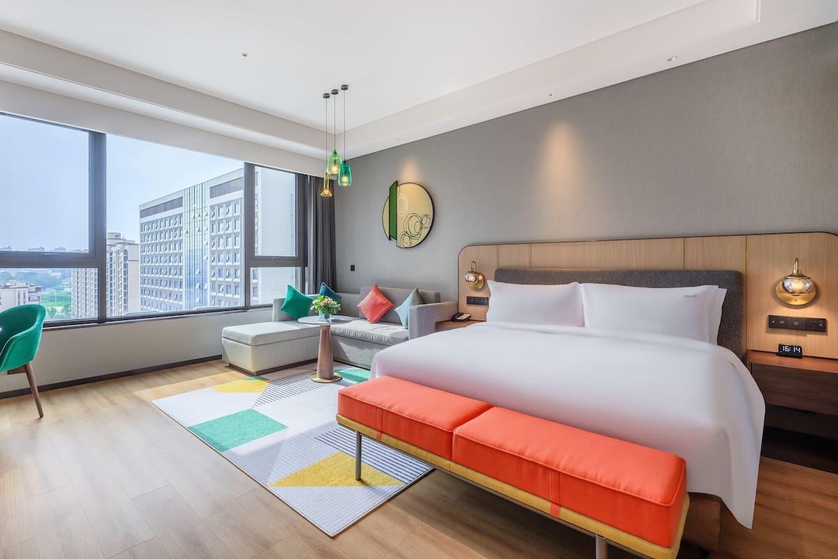 Une chambre d’hôtel moderne, dotée d’un grand lit, d’un bureau et d’un canapé. La fenêtre offre une vue sur de hauts immeubles.