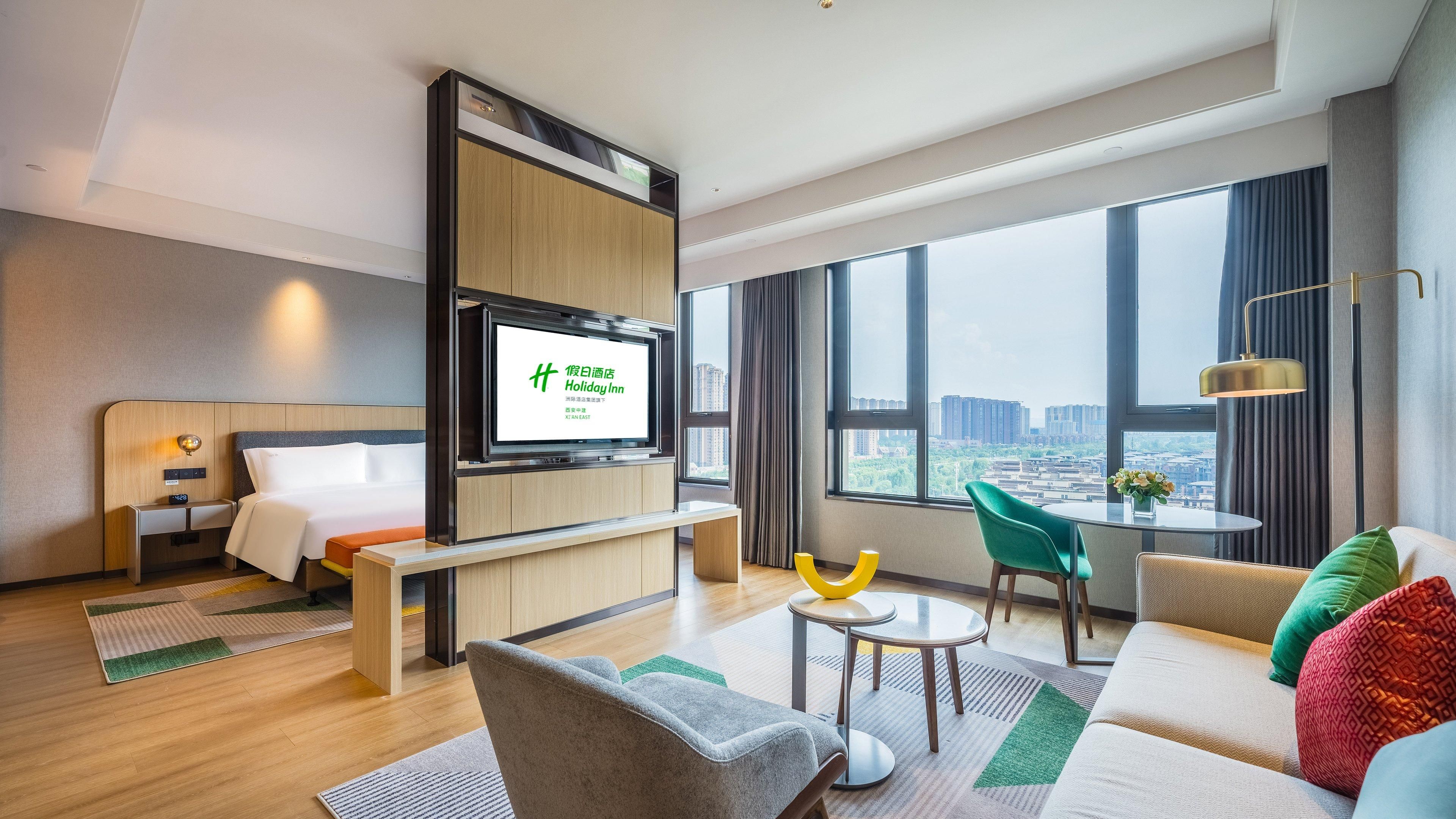 A hotel room with a bed, a TV mounted on a wooden panel, a sofa, a coffee table, and a view of the city through large windows.