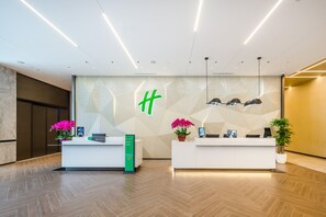 A modern reception area with two white reception desks, a geometric wall design, and a green logo. - Holiday Inn Xian East (Xi'an)