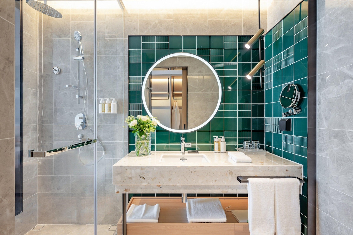 A modern bathroom with a glass-enclosed shower, a round mirror, and a vanity with a sink and towel rack.