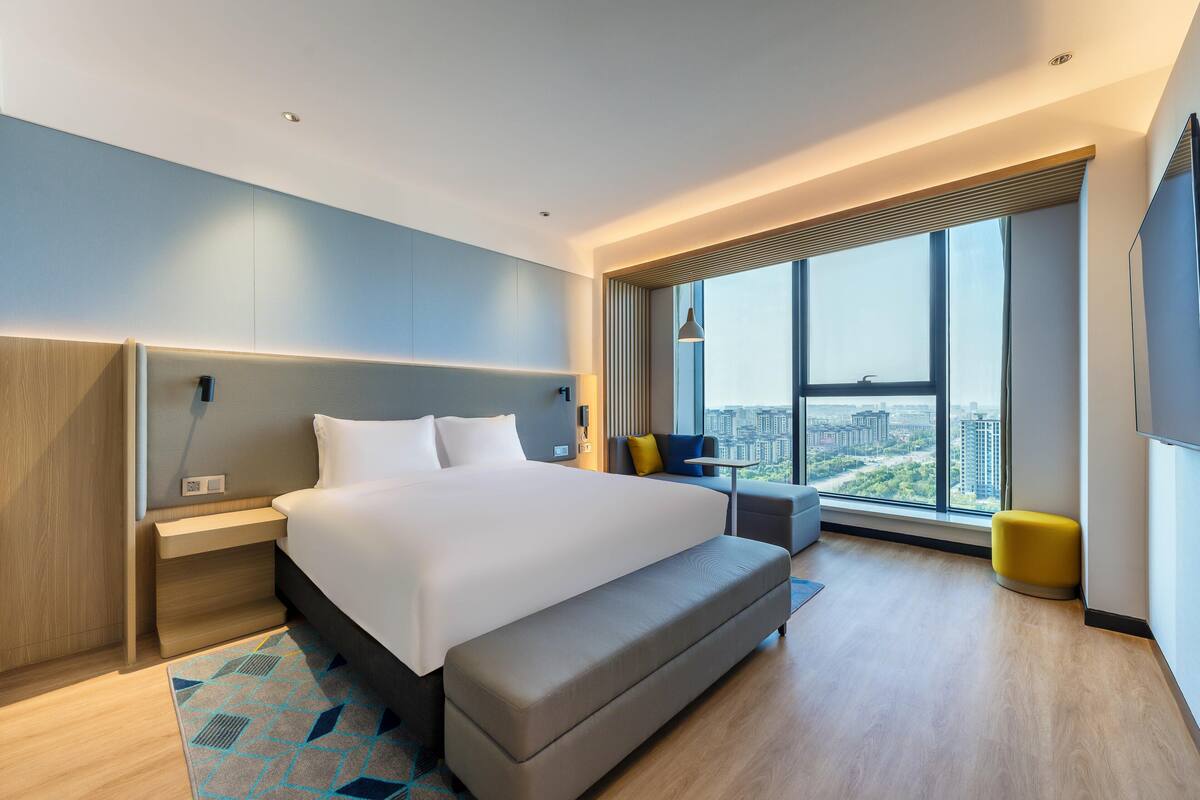A modern hotel room with a large bed, a seating area, a view of the city, and a large window.