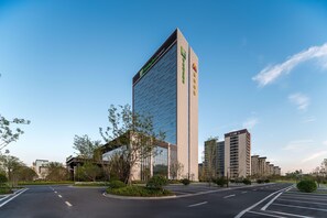 A modern hotel with a prominent glass facade, a parking area, and surrounding greenery.