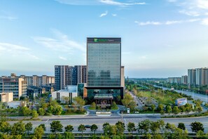 A modern Holiday Inn Express hotel with a glass facade, surrounded by urban buildings and greenery. - Holiday Inn Express Zhoukou Dancheng An Ihg Hotel (Zhoukou)