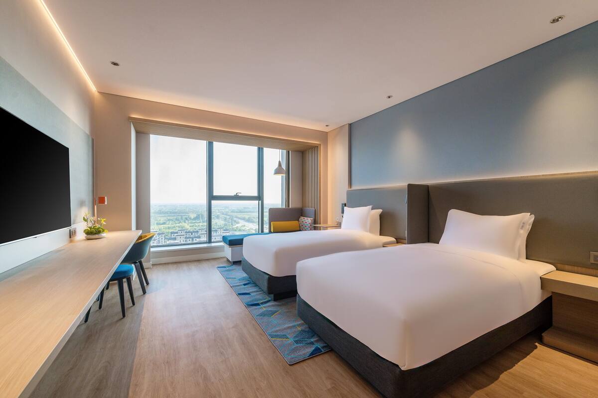 A modern hotel room with two beds, a desk, and a large window offering a city view.
