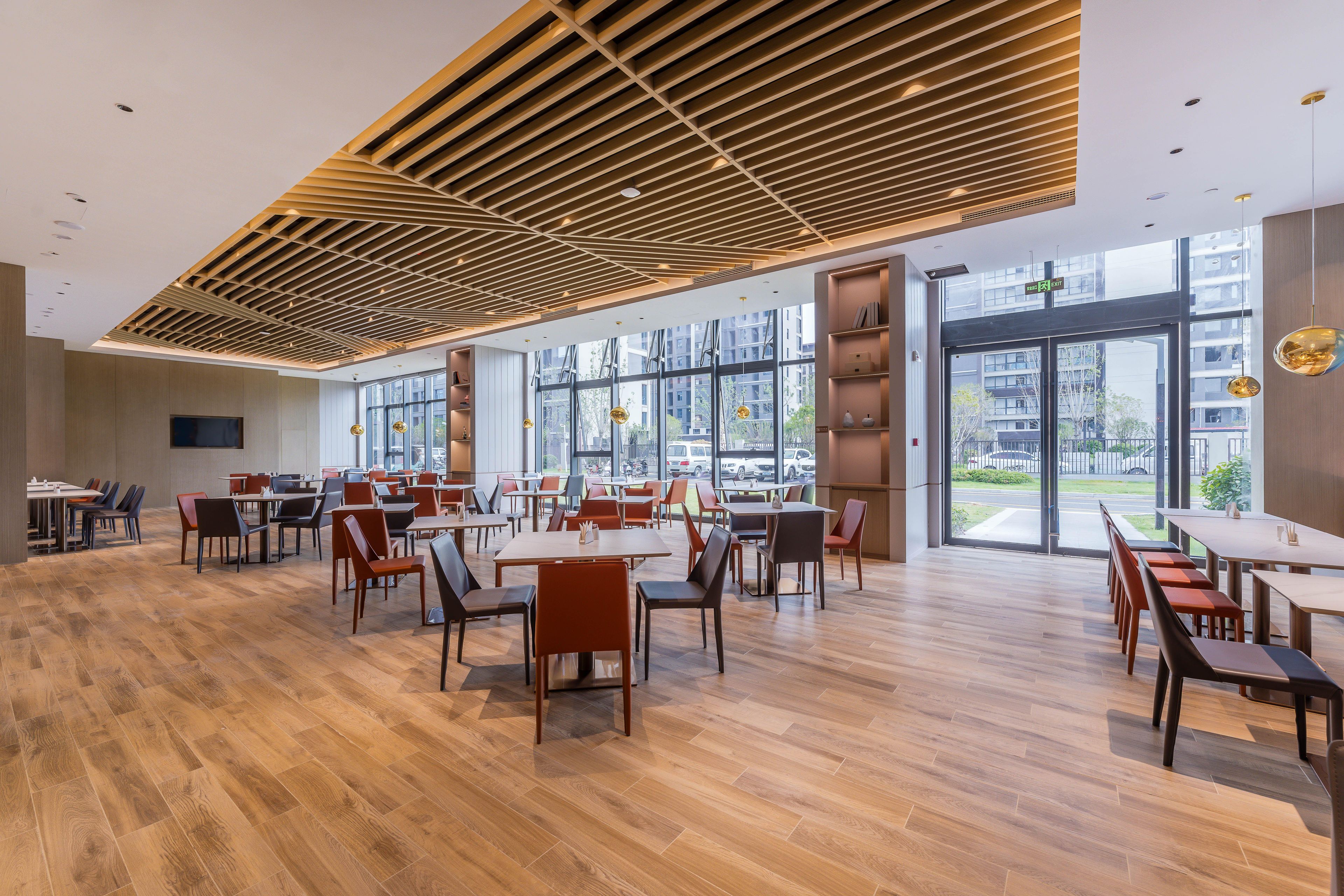 A spacious dining area with wooden flooring and modern furniture, large windows offering views of the city, and a ceiling with a geometric pattern.