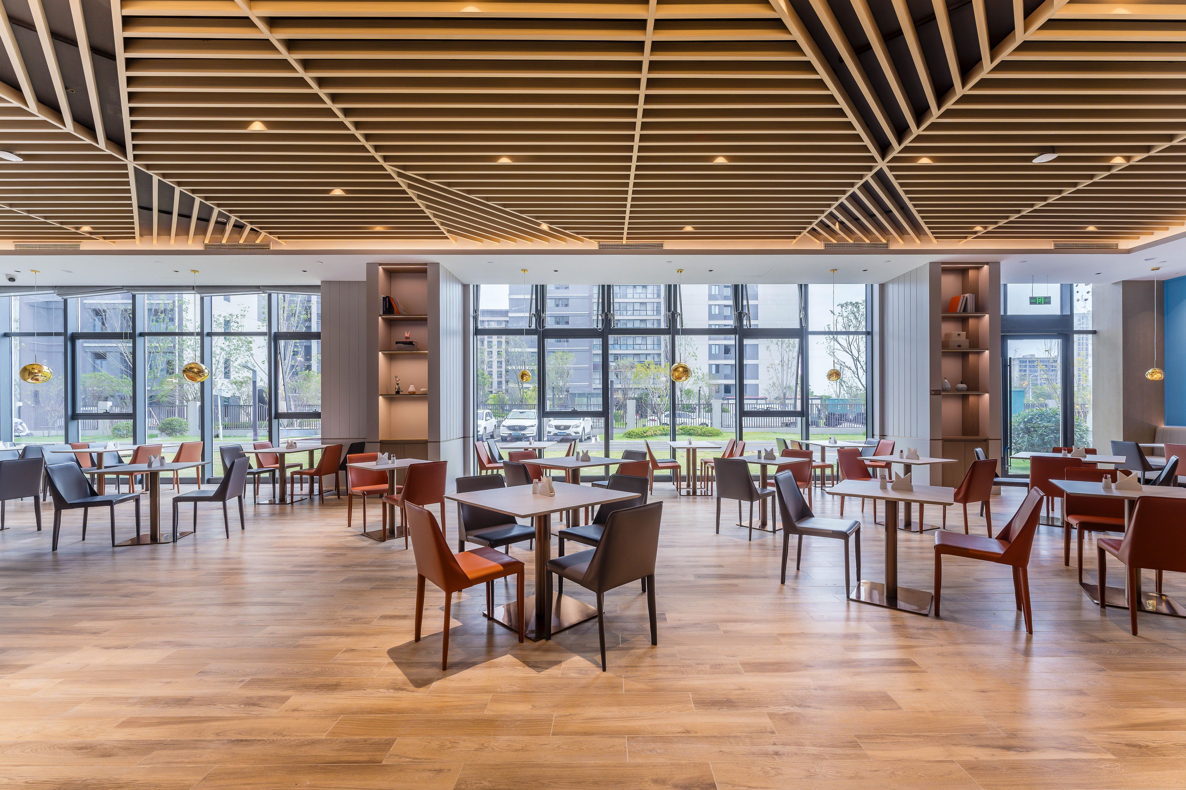 A spacious indoor area with wooden flooring, tables, and chairs arranged for dining. Large windows provide views of the outside.