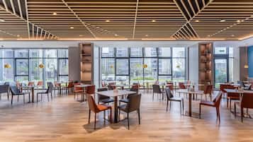 A spacious indoor area with wooden flooring, tables, and chairs arranged for dining. Large windows provide views of the outside.