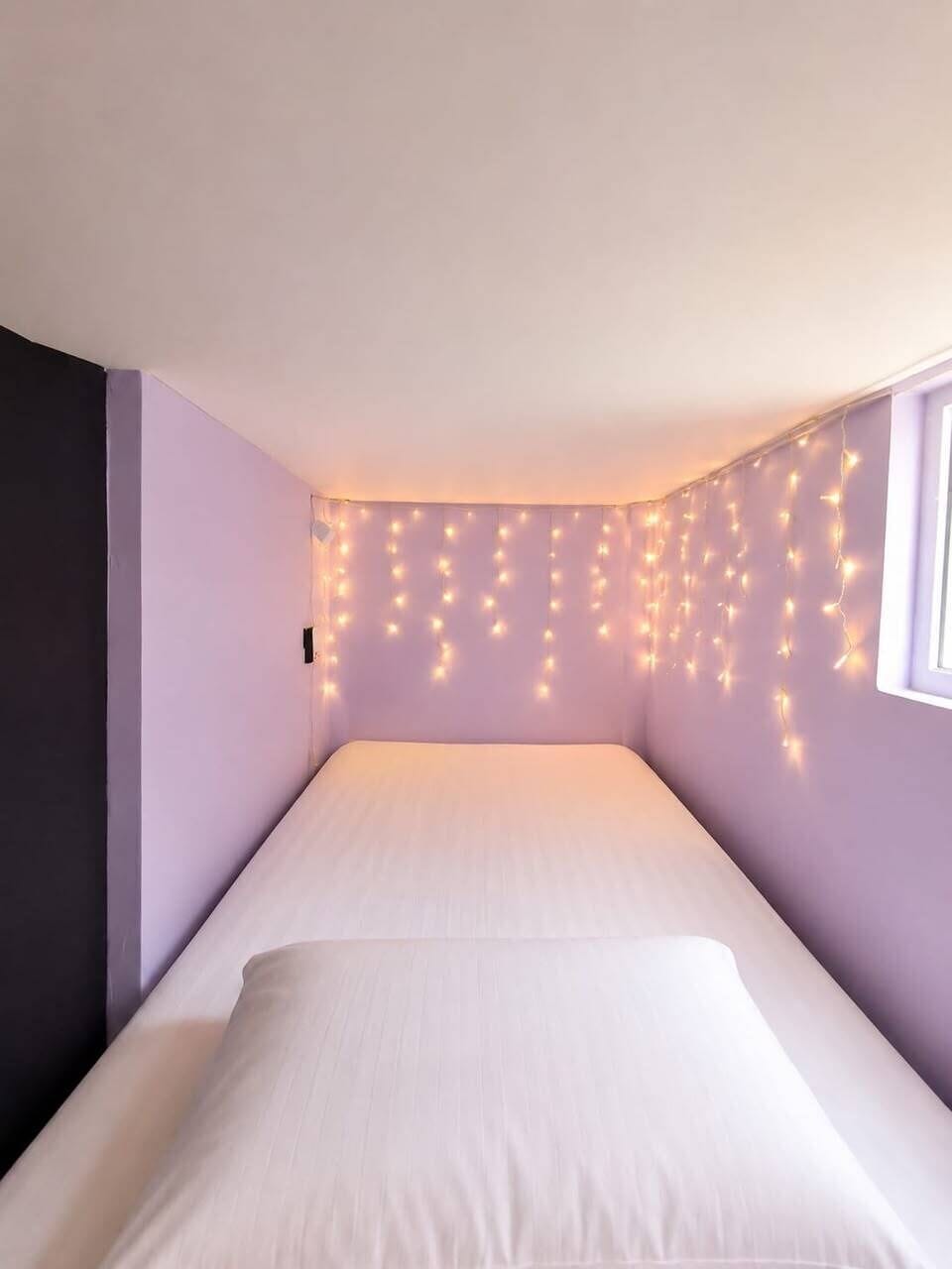 A small, dimly lit room with a single bed, a window on the right, and string lights on the wall.