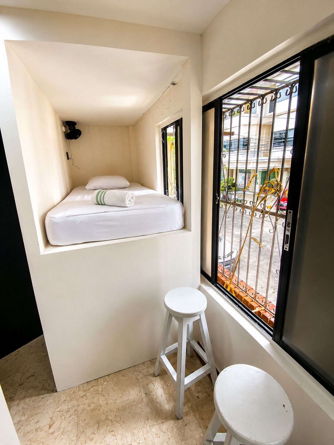 A small, compact room with a bed, a stool, and a window with a metal gate.