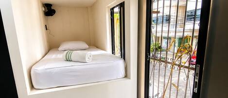 A small, compact room with a bed, a stool, and a window with a metal gate.