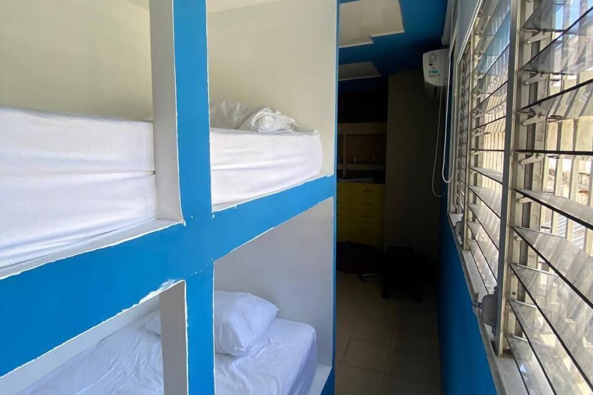 Un couloir étroit avec des lits superposés, une fenêtre grillagée et une petite pièce avec un bureau et une chaise.