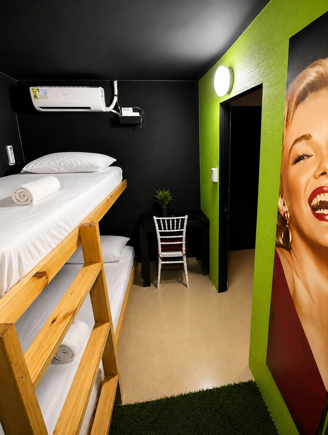 Een kamer met een stapelbed, een bureau, een stoel, een kleine plant en een luchtverkoeler aan de muur.
