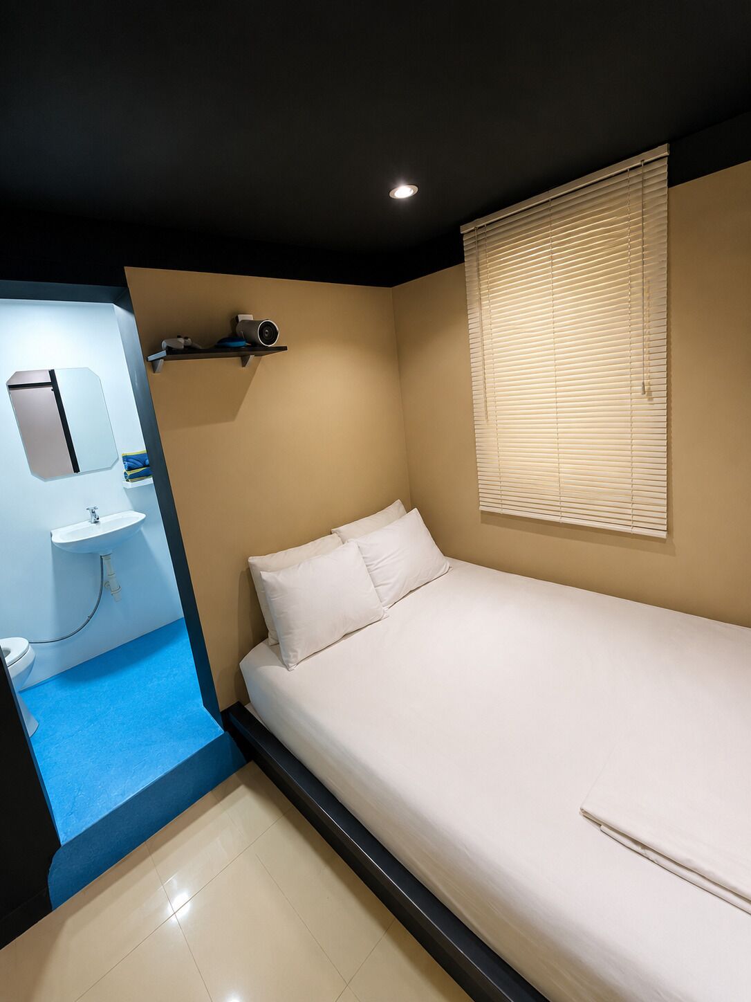 Un dormitorio con una cama individual, una mesita de noche, una ventana con persianas, un espejo y un baño con lavabo e inodoro.