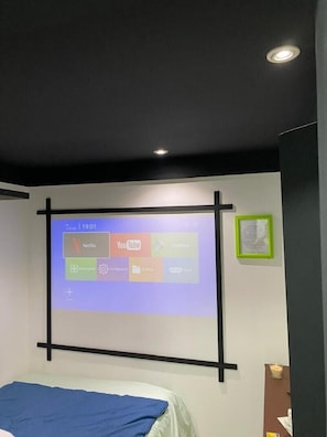 Una pantalla de proyección mostrando iconos de aplicaciones, una cama con una colcha azul y un diploma enmarcado en la pared.