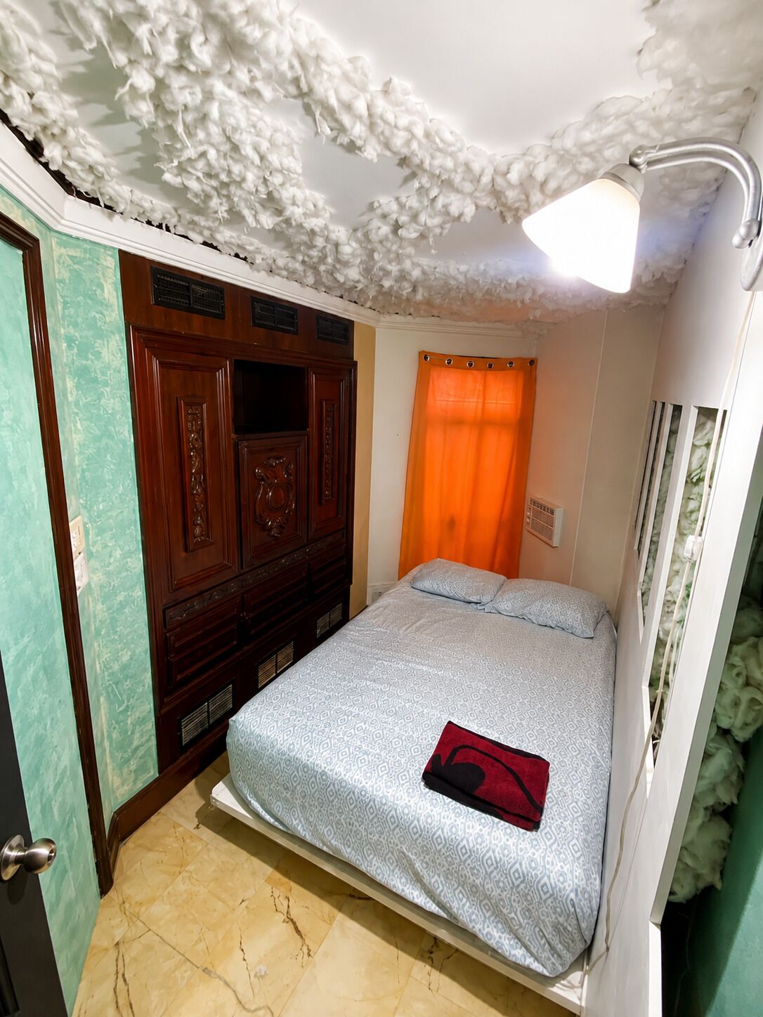 A bedroom with a bed, a wooden wardrobe, a window with an orange curtain, and a ceiling with a fluffy texture.