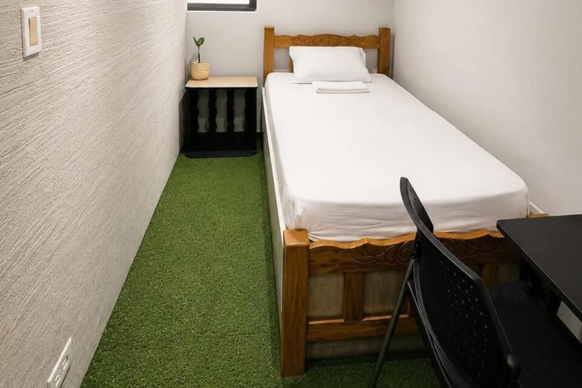 Une petite chambre avec un lit simple, une tête de lit en bois, une petite table de chevet, une chaise et une fenêtre avec des stores.