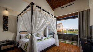 A four-poster bed with white drapes, a wooden floor, a nightstand, a wall-mounted artwork, and a view of a building through a window. - Mokko Suites Batubelig (Keroboken Kelod)