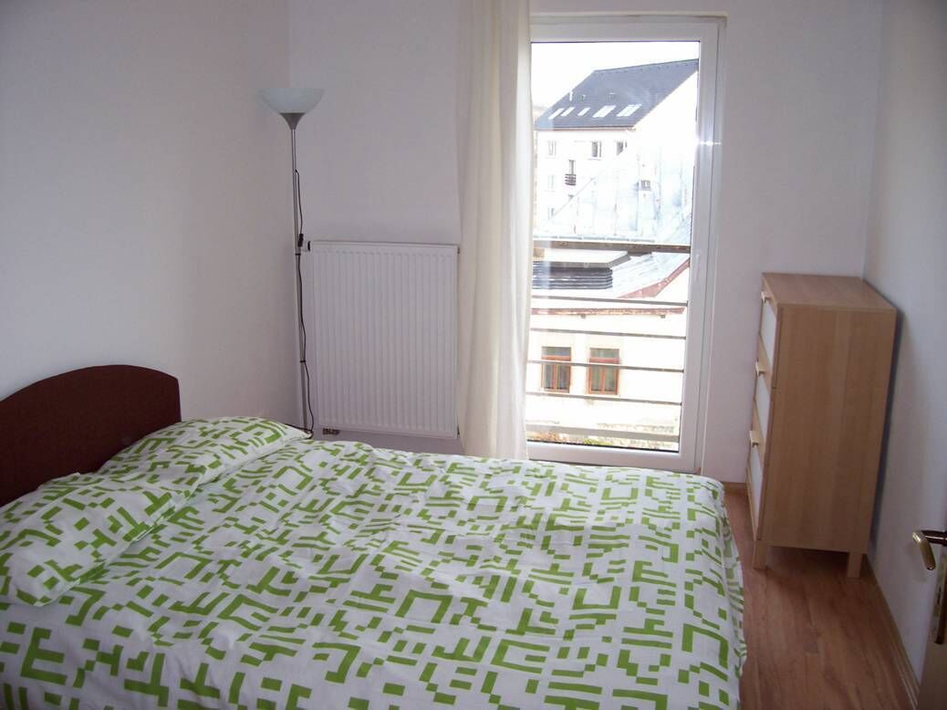 A bedroom with a bed, a radiator, a wooden cabinet, and a window with a view of buildings.