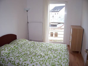 A bedroom with a bed, a radiator, a wooden cabinet, and a window with a view of buildings.
