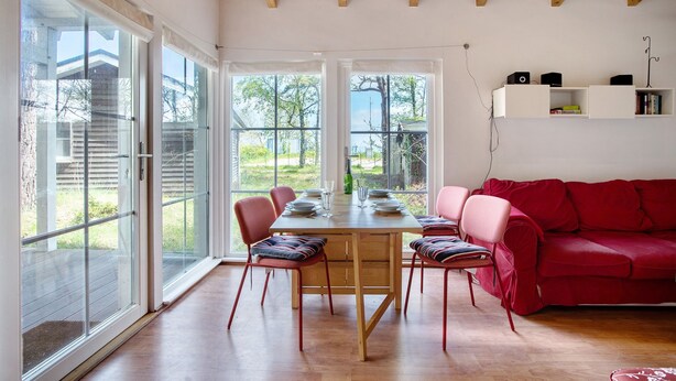 Un comedor con una mesa y sillas de madera, un sofá rojo y ventanas grandes con vistas a la vegetación.