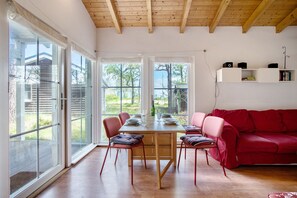 Un coin repas avec une table et des chaises en bois, un canapé rouge et de grandes fenêtres donnant sur un paysage verdoyant.