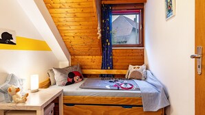 A cozy room with a wooden ceiling, a bed with Mickey Mouse bedding, a small desk with a lamp, and a window with curtains.