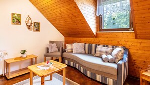 A cozy room with a sloped ceiling, a sofa, a chair, a small table, and a window with a view of trees.