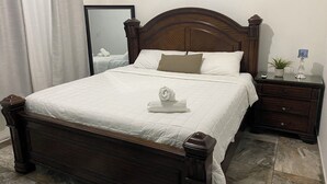 A neatly made bed with a white duvet, a wooden headboard, and a bedside table with a lamp and a small plant. - Pompeii Apartment (Aguada)