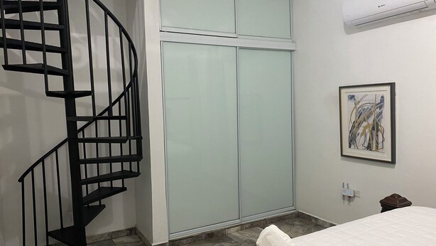 A bedroom with a spiral staircase, a sliding glass door closet, a framed abstract painting, and a neatly made bed with rolled towels. - Pompeii Apartment (Aguada)