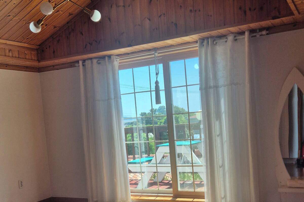 A room with wooden flooring, a large window with curtains, and a ceiling with wooden beams and hanging lights.