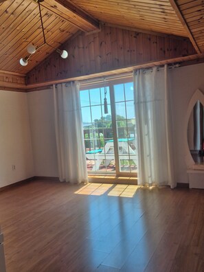 Une piĂšce dotĂ©e dâun plancher en bois, dâune grande fenĂȘtre avec des rideaux et dâun plafond ornĂ© de poutres en bois et de luminaires suspendus.