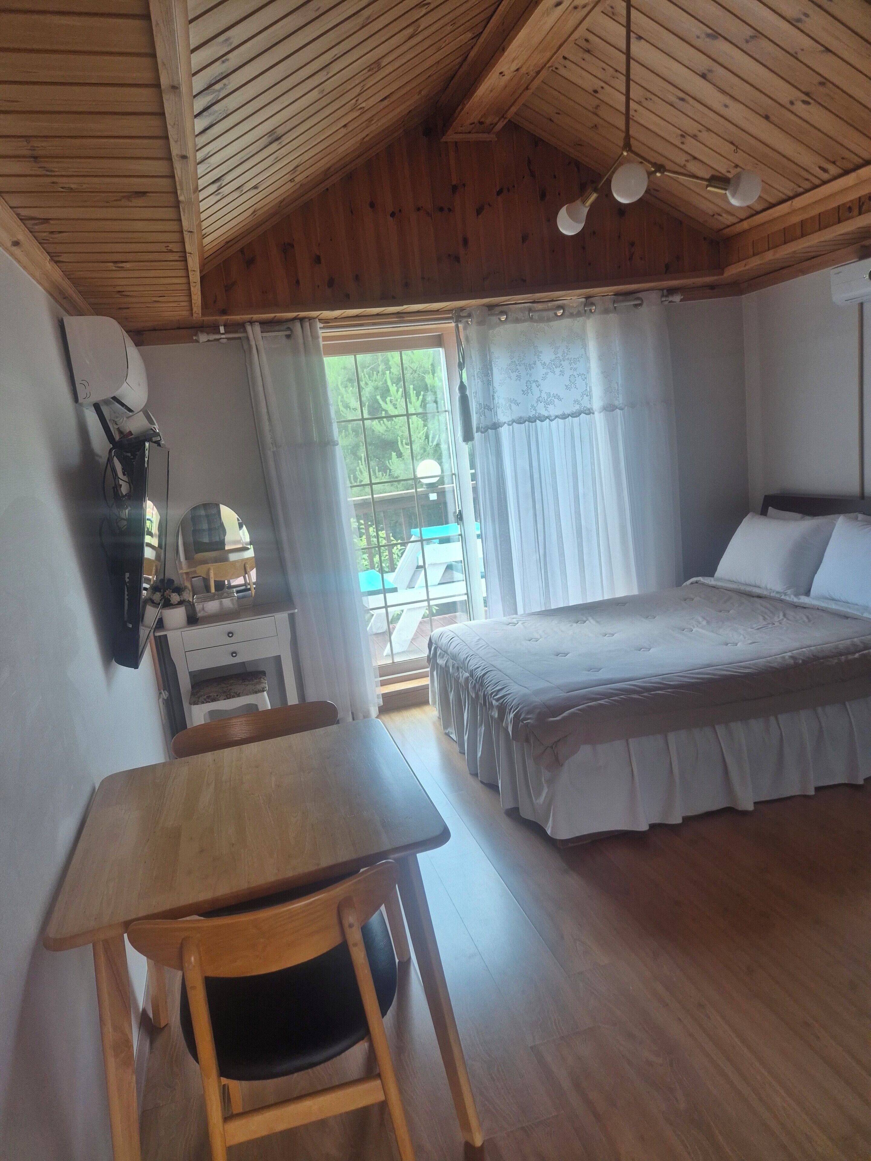 Ein Schlafzimmer mit Holzbalkendecke, einem Bett mit weißer Bettwäsche, einem Schreibtisch mit Stuhl und einem Fenster mit Vorhängen.