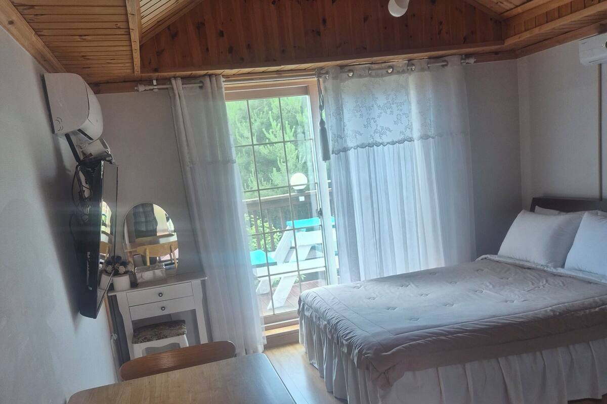A bedroom with a wooden ceiling, a bed with white bedding, a desk with a chair, and a window with curtains.