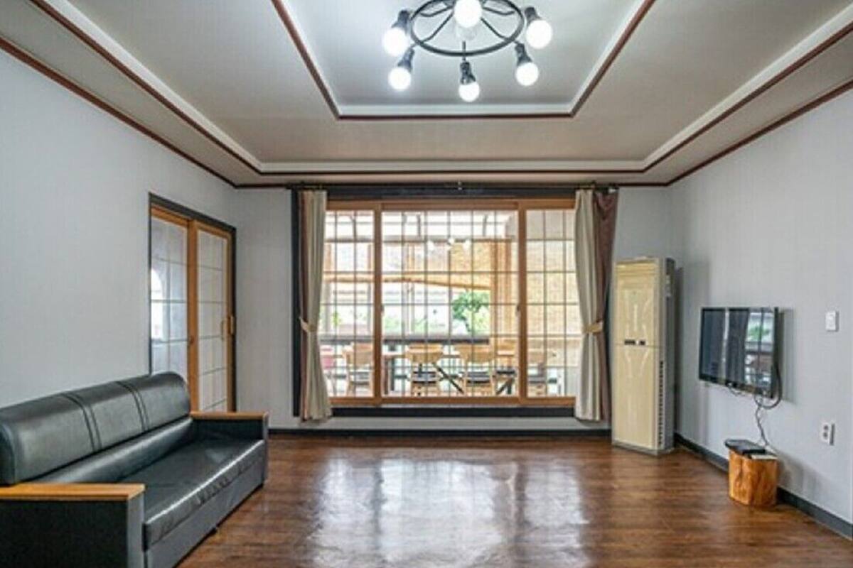 A spacious room with a large window, a black leather sofa, a wooden floor, and a ceiling light fixture.