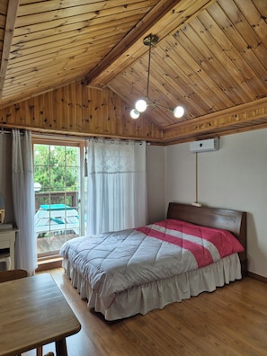 Un dormitorio con techo de madera, una cama con colcha rosa y blanca, una mesa de madera y una ventana con cortinas.